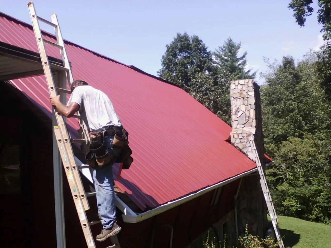 Roofer on a ladder beside a red metal roof and stone chimney — Soffit & Fascia Repair by Roofing Brothers in Fort Riley