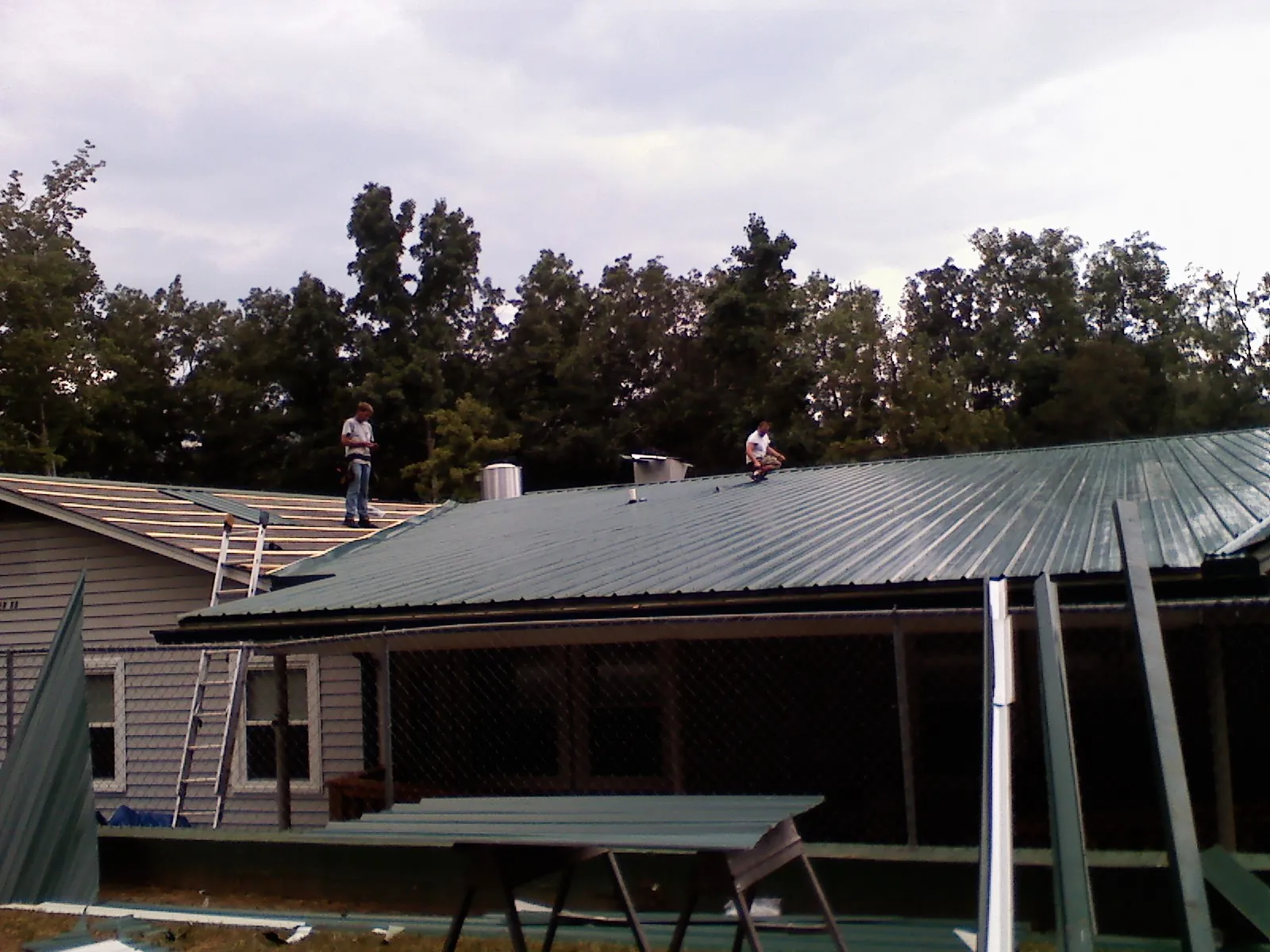 Two roofers installing green metal panels above a covered porch in Fort Riley